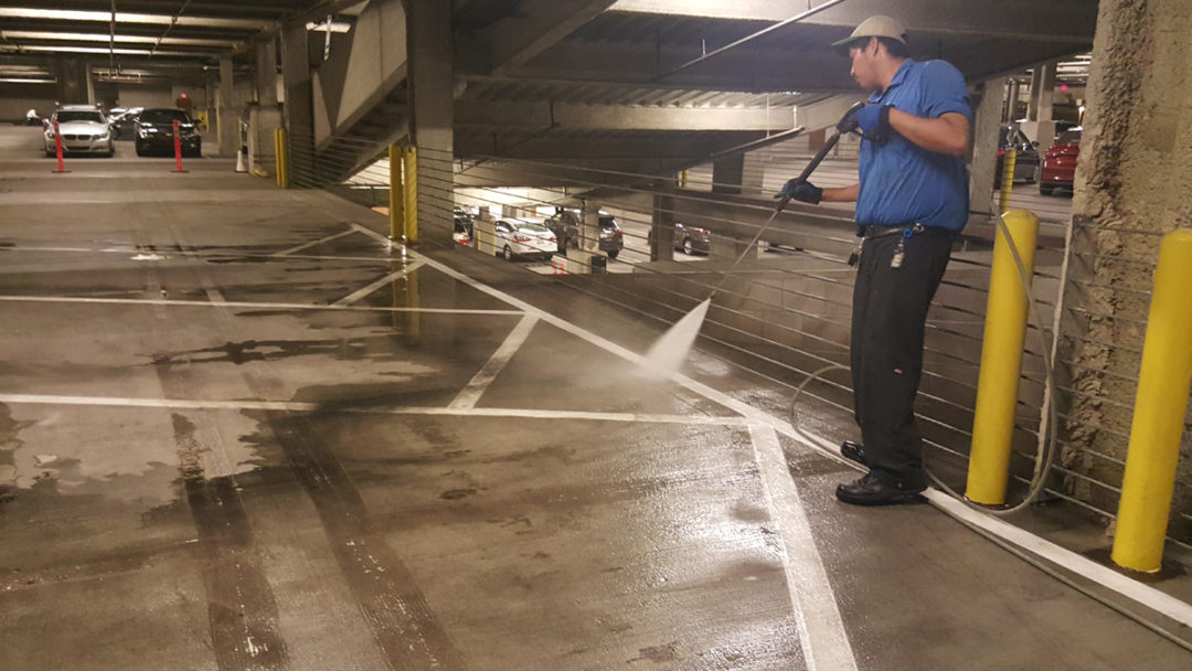 Man in a blue shirt using a pressure washer to clean concrete floor in a parking garage.